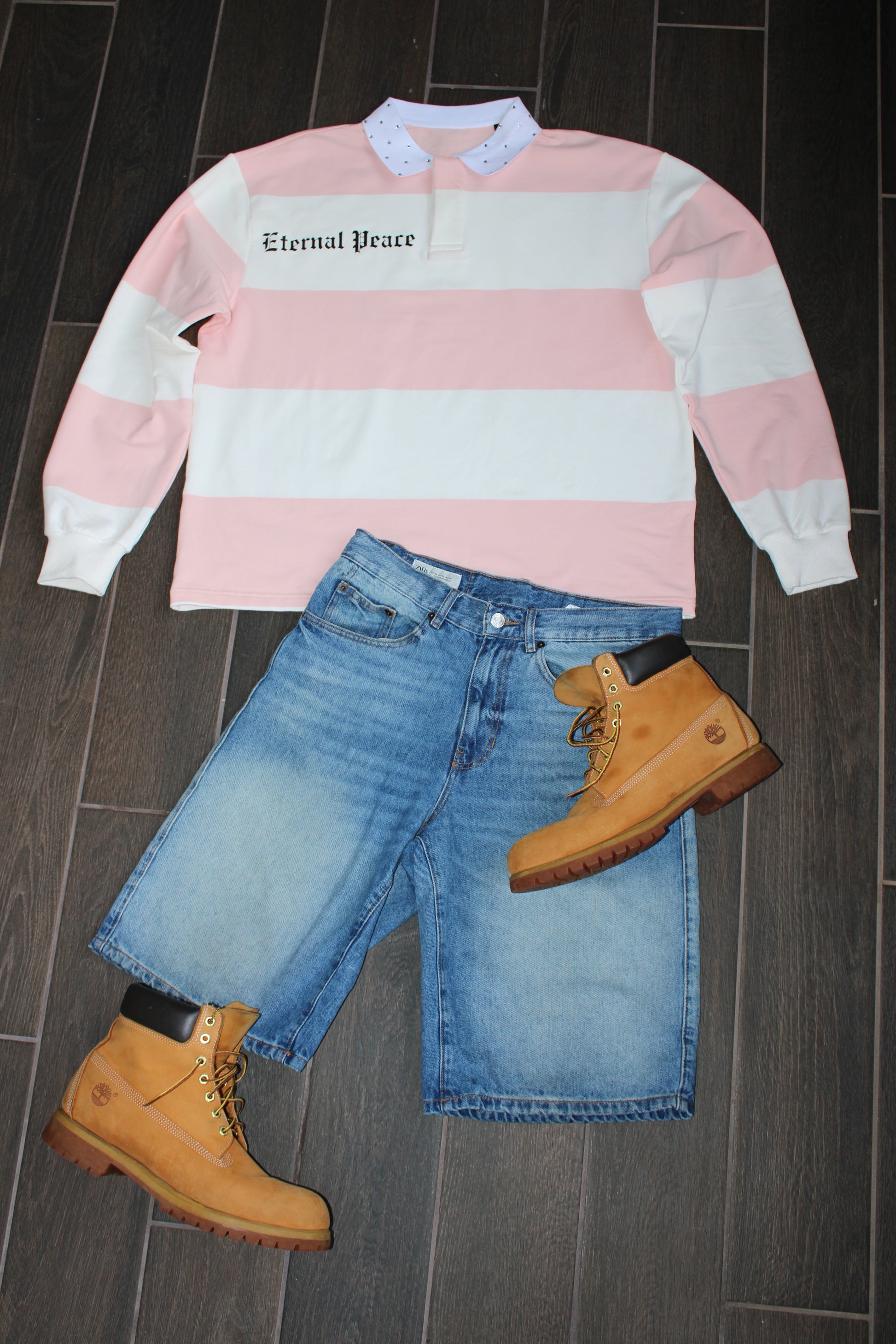 Pink and white striped sweatshirt with 'Eternal Peace' text, blue jeans, and brown boots on a dark wooden floor.